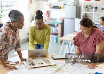 Stock image of 3 women looking at blueprints, a laptop, and a house model