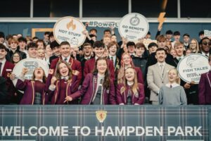 A group of school pupils stand behind a banner that reads 'Welcome to Hampden Park', holding Powering Futures related signs.
