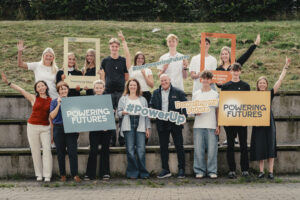 A group of Powering Futures participants and facilitators hold props such as frames and signs, some have raised hands.
